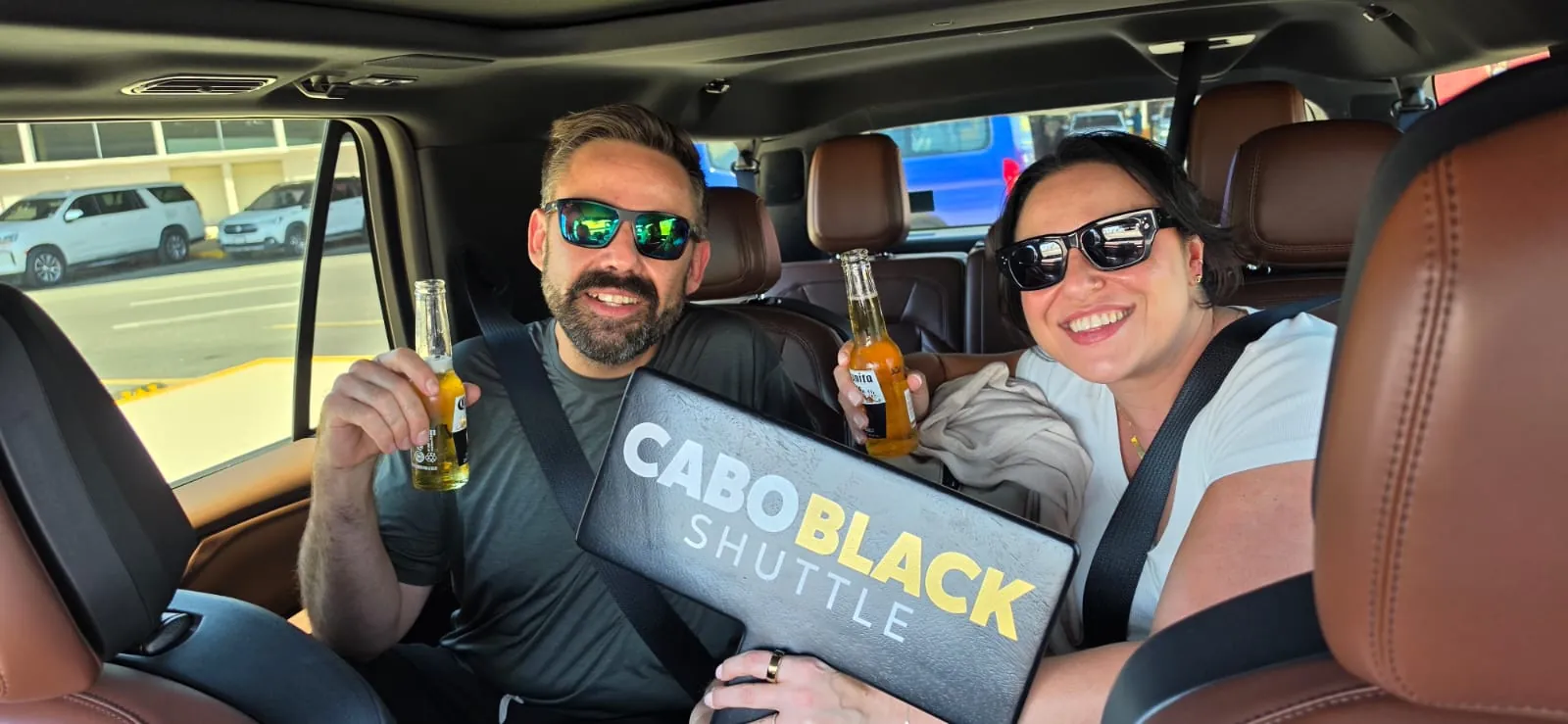 Couple holding cold beers and a Cabo Black Shuttle sign in the back seat of a private SUV at SJD airport