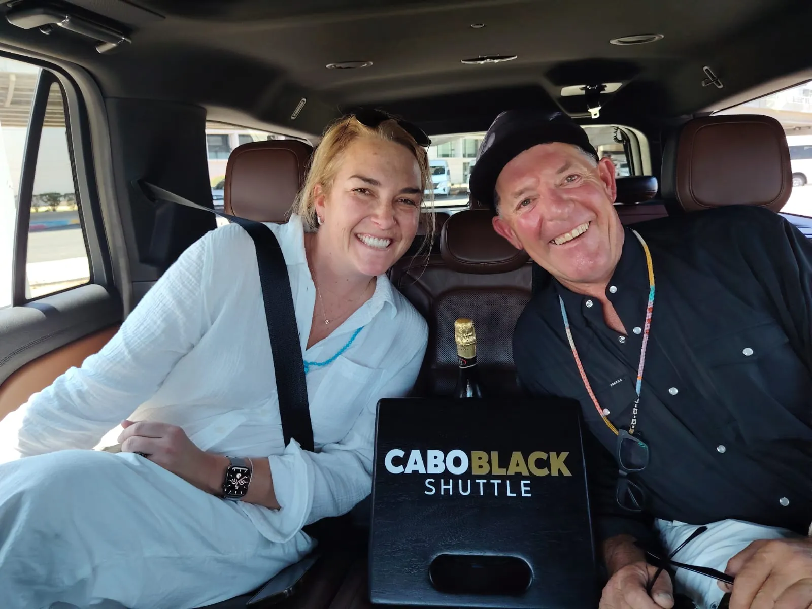 Two guests in the leather back seat of a private SUV with a bottle of sparkling wine and a Cabo Black Shuttle sign